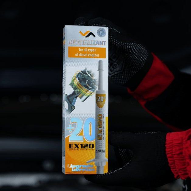 A close-up of a person wearing a black-and-red mechanic's suit, holding a box of a revitalizing product for vehicles.