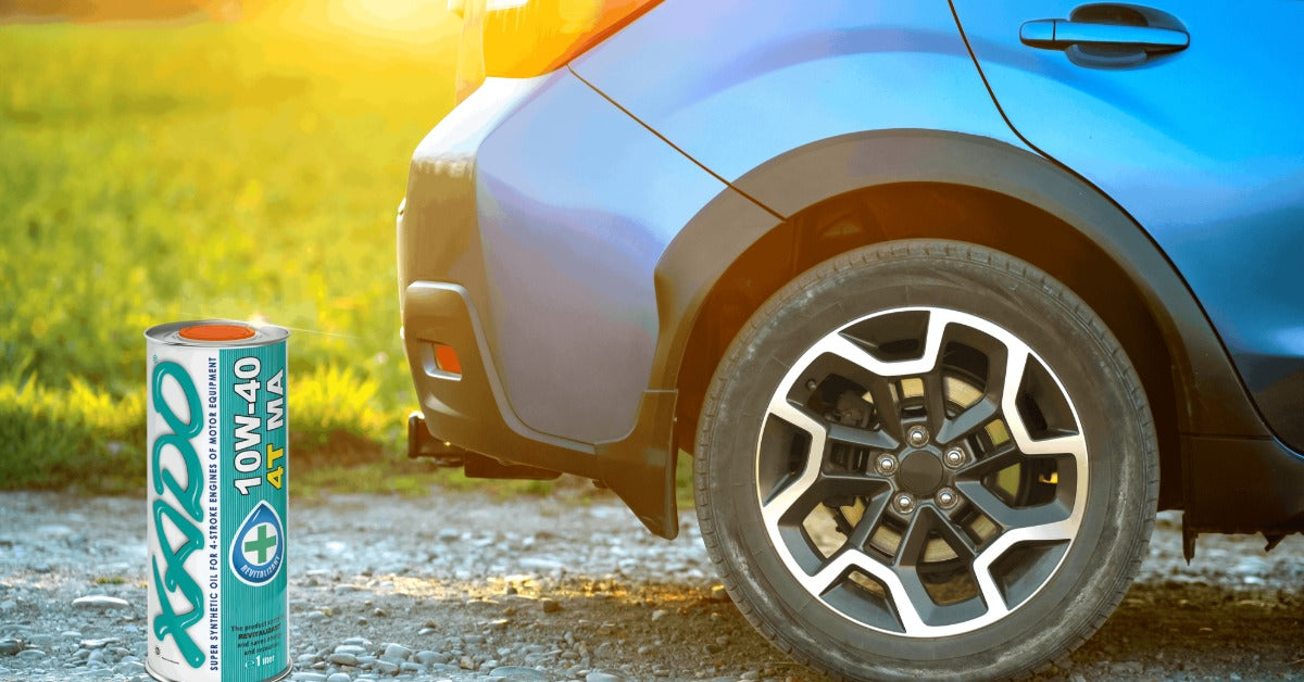 A low-angle view of a car parked on a gravel road near a grassy field, with a canister of XADO product placed near it.