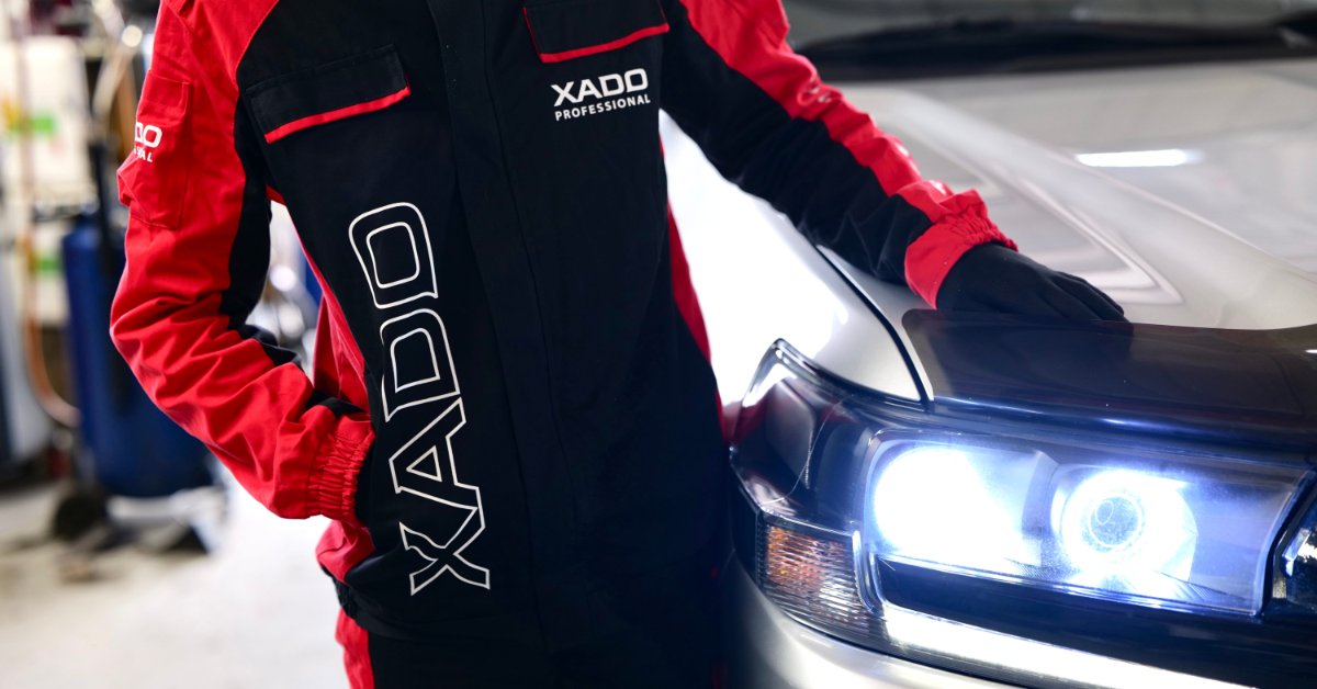 A person wearing a red-and-black mechanic suit stands next to a white vehicle with the headlights turned on.