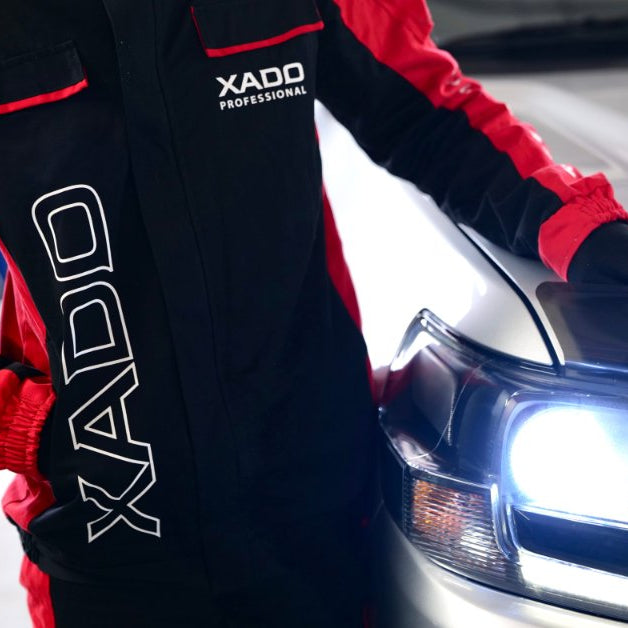 A person wearing a red-and-black mechanic suit stands next to a white vehicle with the headlights turned on.