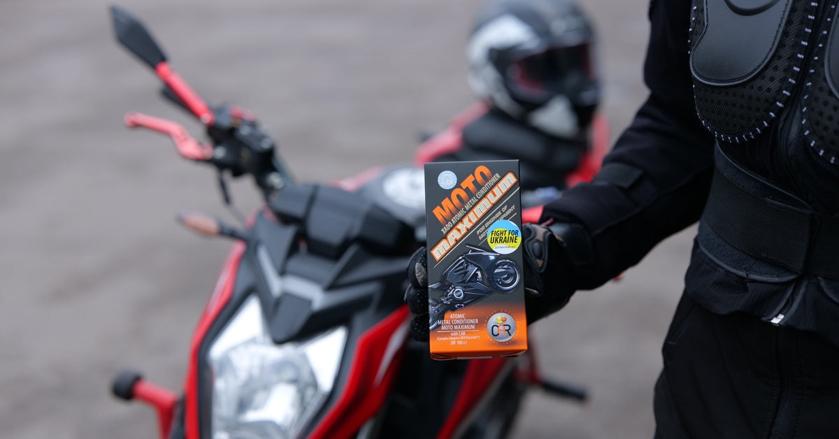 A person in black motorcycle gear holding a box of motor treatment beside a motorcycle with a helmet behind it.