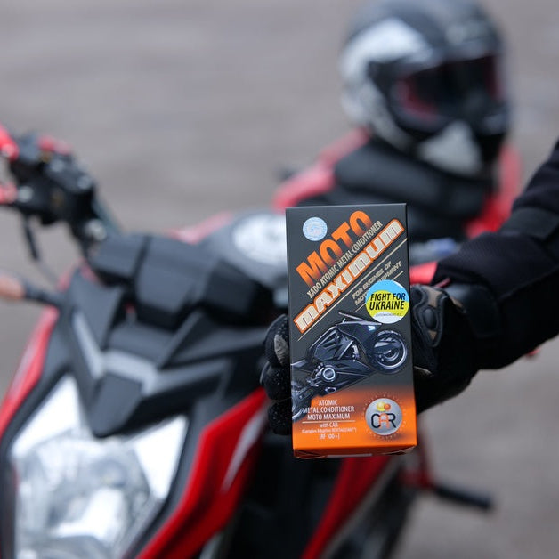 A person in black motorcycle gear holding a box of motor treatment beside a motorcycle with a helmet behind it.