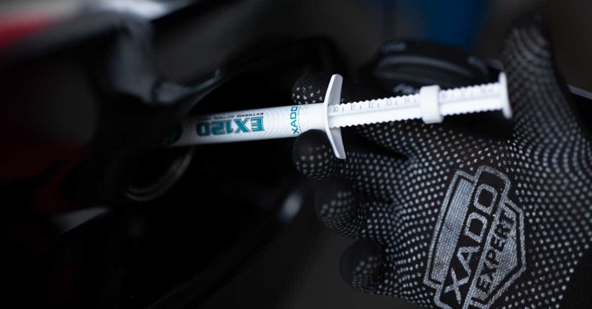 A gloved hand holding a white product syringe near a dark vehicle area with the syringe label facing outward.