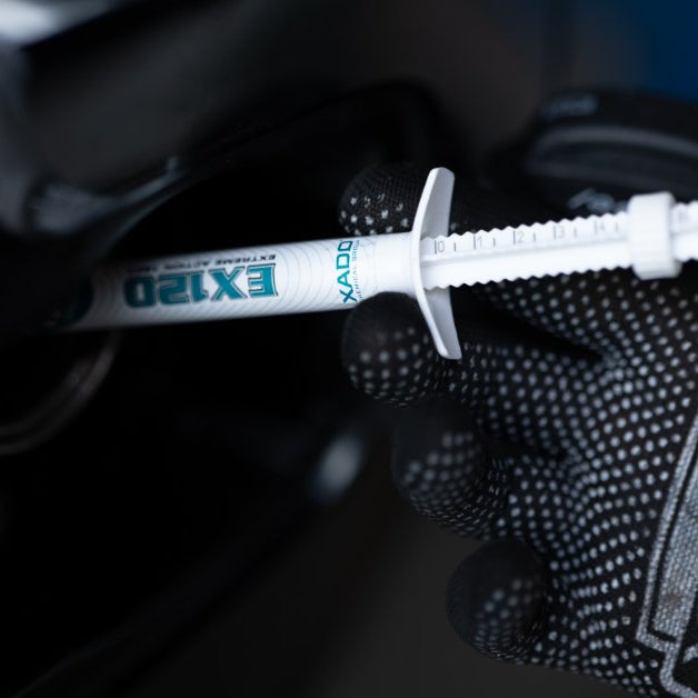 A gloved hand holding a white product syringe near a dark vehicle area with the syringe label facing outward.