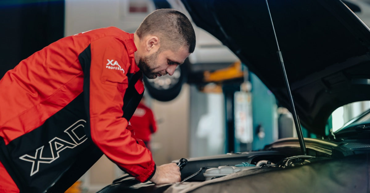 A man in a red and black jacket leaning over an open car hood while working in a vehicle repair garage.