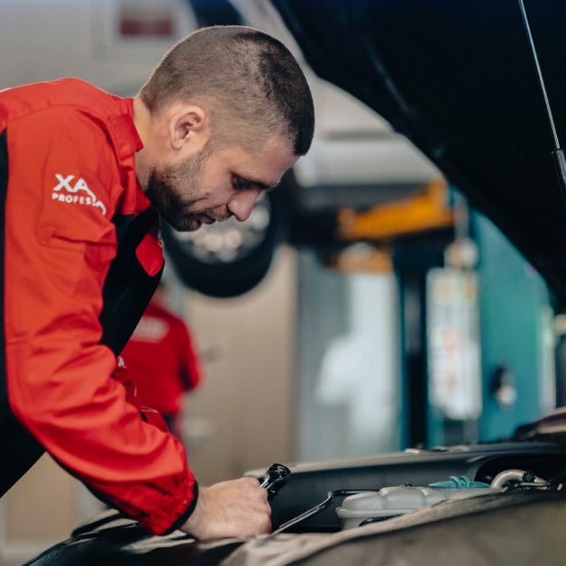 A man in a red and black jacket leaning over an open car hood while working in a vehicle repair garage.